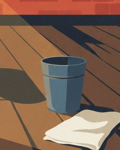 Un seau en métal bleu et une serviette blanche pliée sur le parquet, dans l'ombre des fenêtres